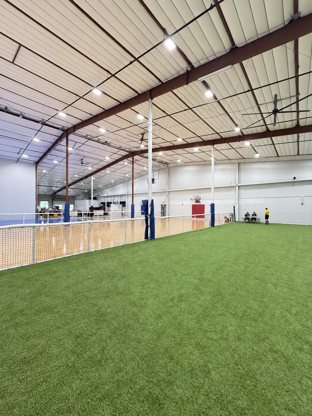 Full indoor turf field with perimeter netting and hard courts visible beyond the divider, set up for tournament and league play