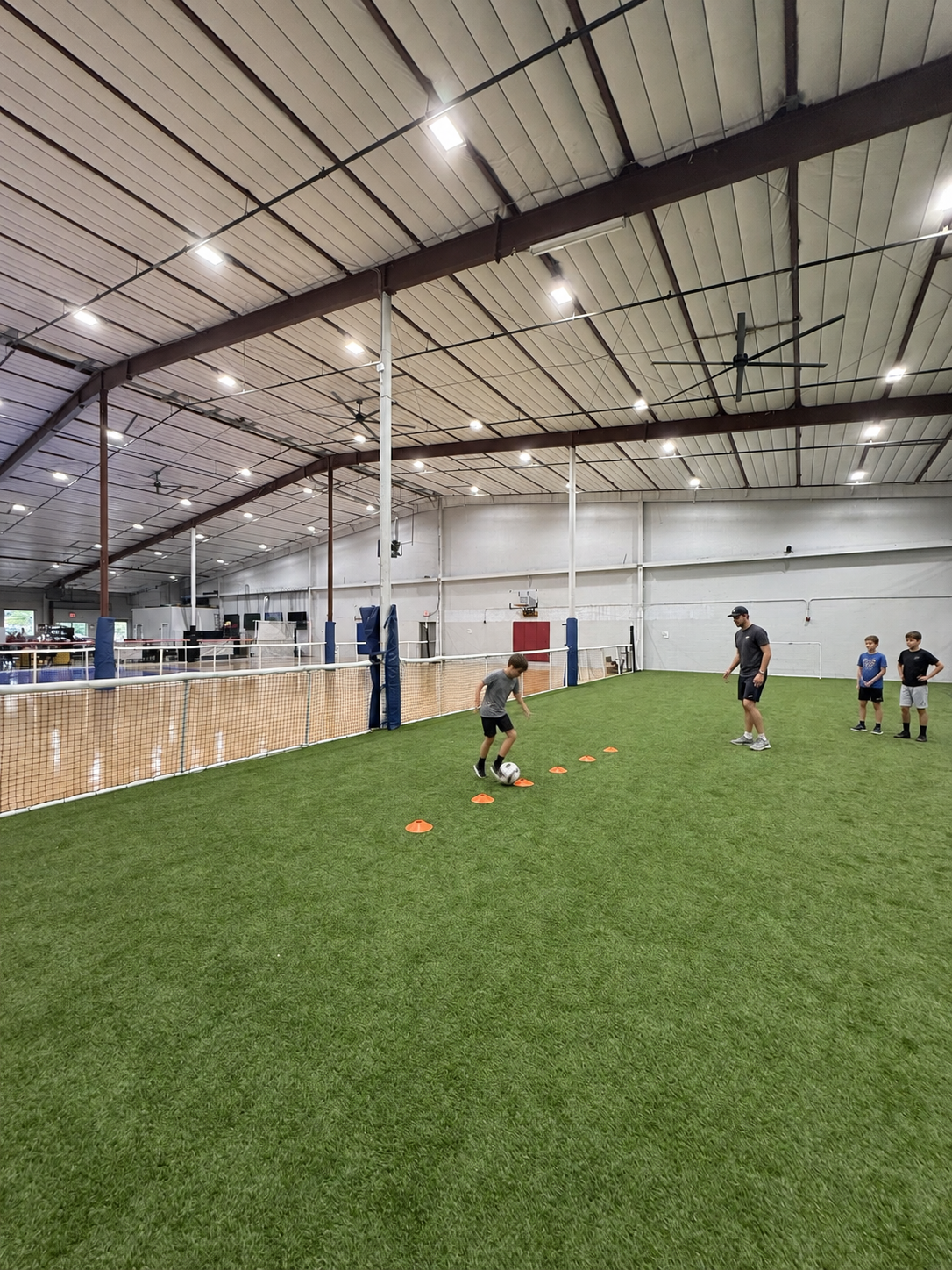 Children practicing soccer dribbling around cones on the indoor turf field with a coach, at PSA Sports Club