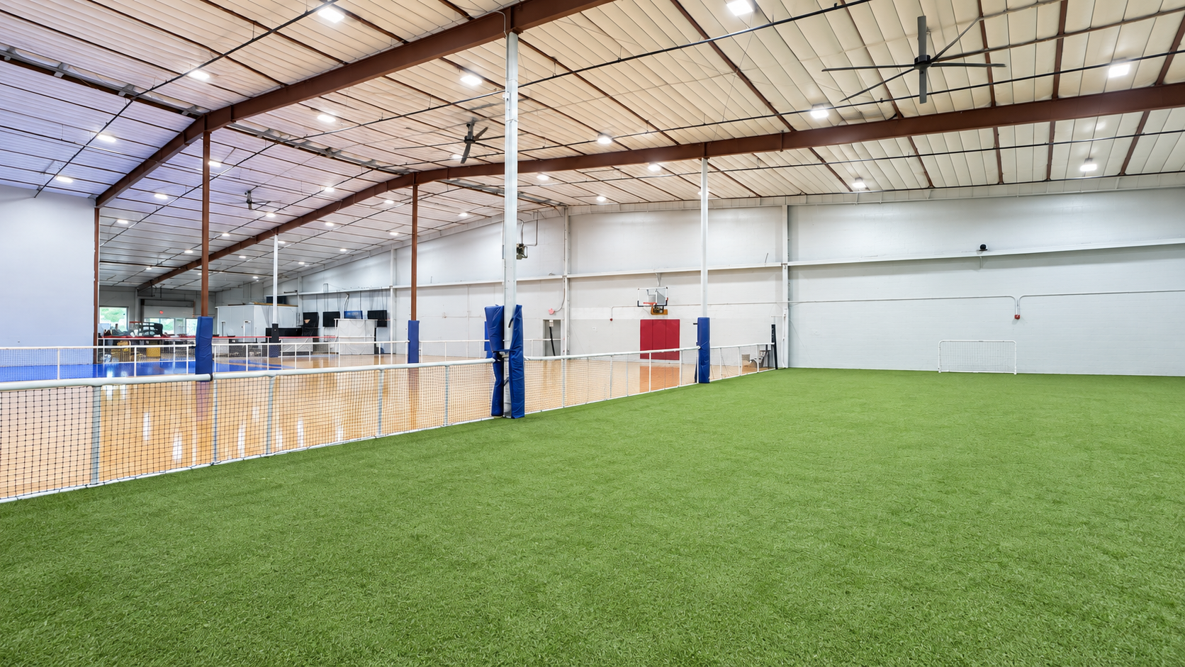 Wide interior view of PSA Sports Club showing the green turf field in the foreground, a white netting divider along the left, wood and blue hard courts beyond, a basketball hoop and red wall padding on the far wall, blue-padded support posts, exposed brown roof trusses and large ceiling fans overhead. No people are present.